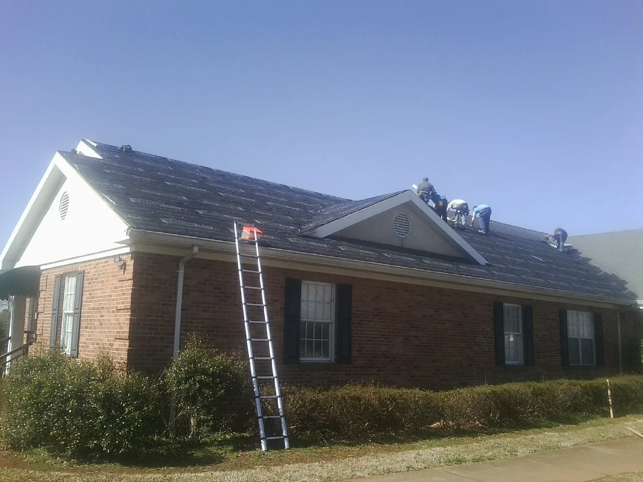 Crew tearing shingles off a brick ranch roof with a ladder set — Emergency Roof Repair work by our crew in New Raymer
