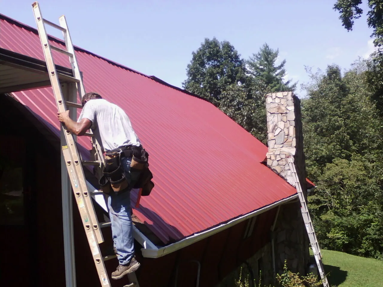 Roofer on a ladder beside a red metal roof and stone chimney — Soffit & Fascia Repair by Roofing Brothers in New Raymer